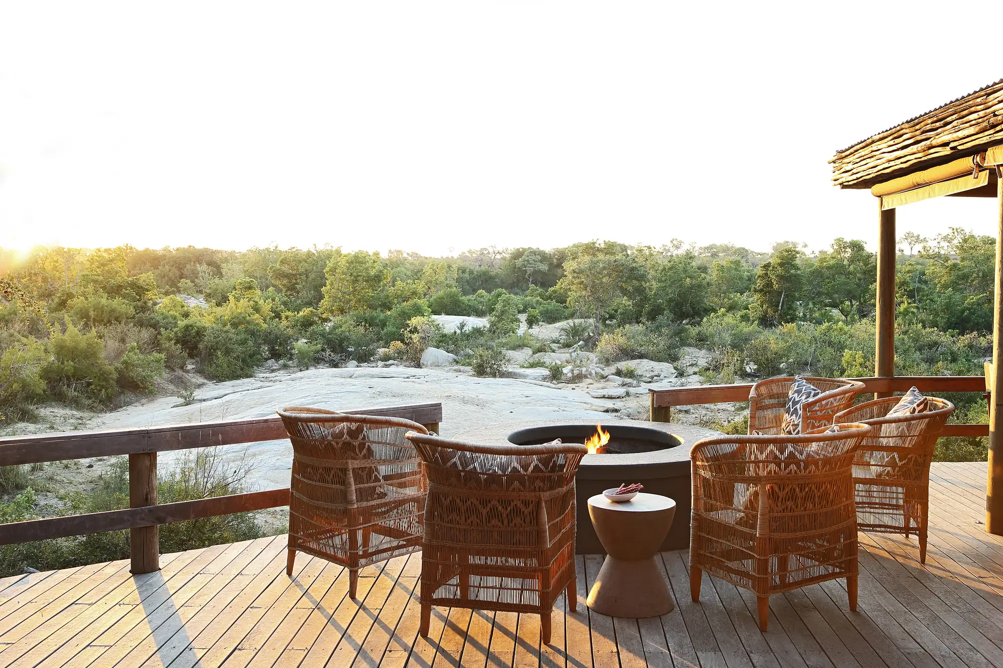 The fire deck at Founders Camp at sunset — five woven armchairs around a low stone fire bowl on a timber deck, looking out over the pale rock bed of the Sand River and the bush beyond.