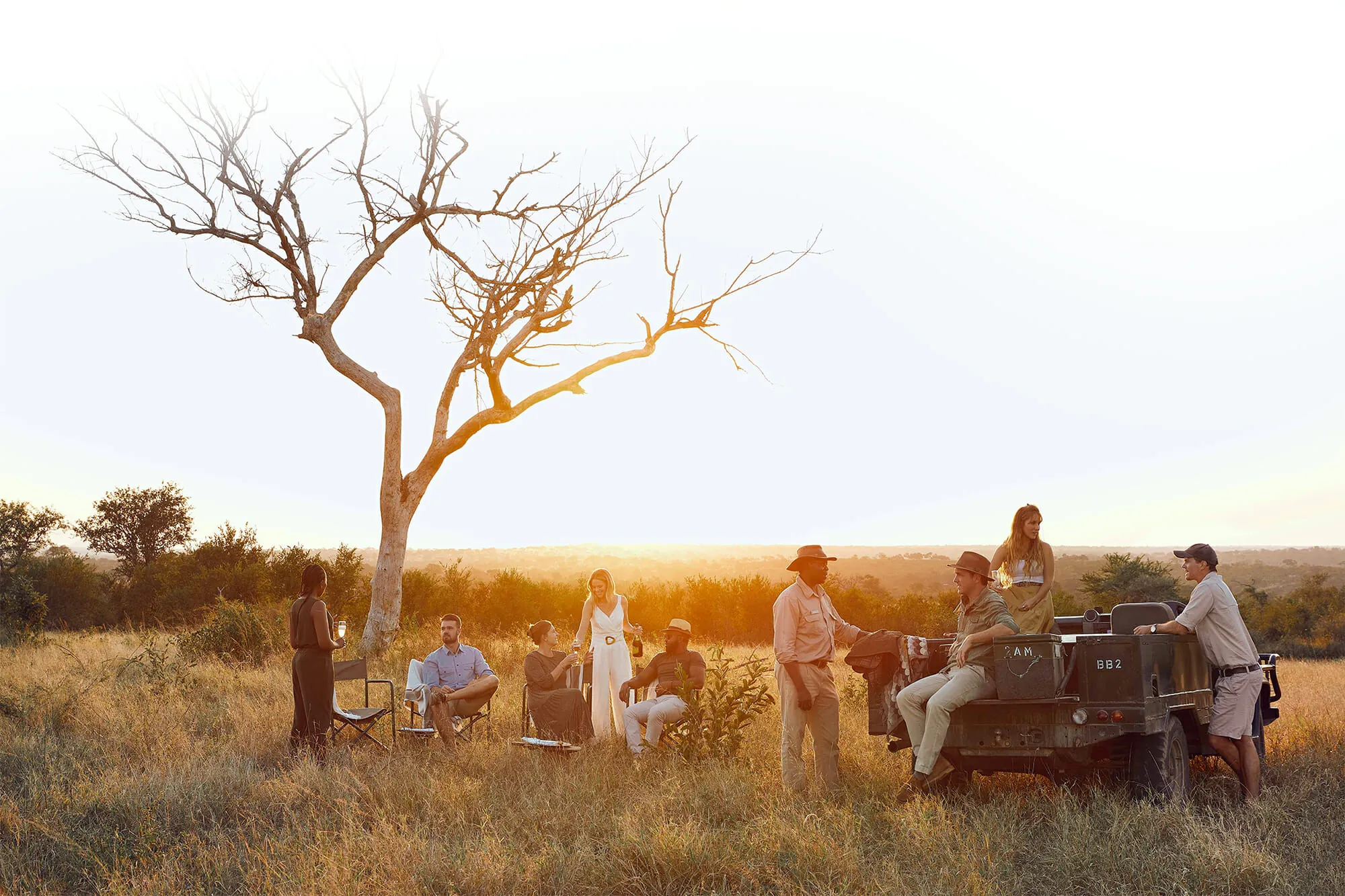 A group of people standing and seated around an open safari vehicle beneath a bare tree in open grassland, with the sun low behind them.