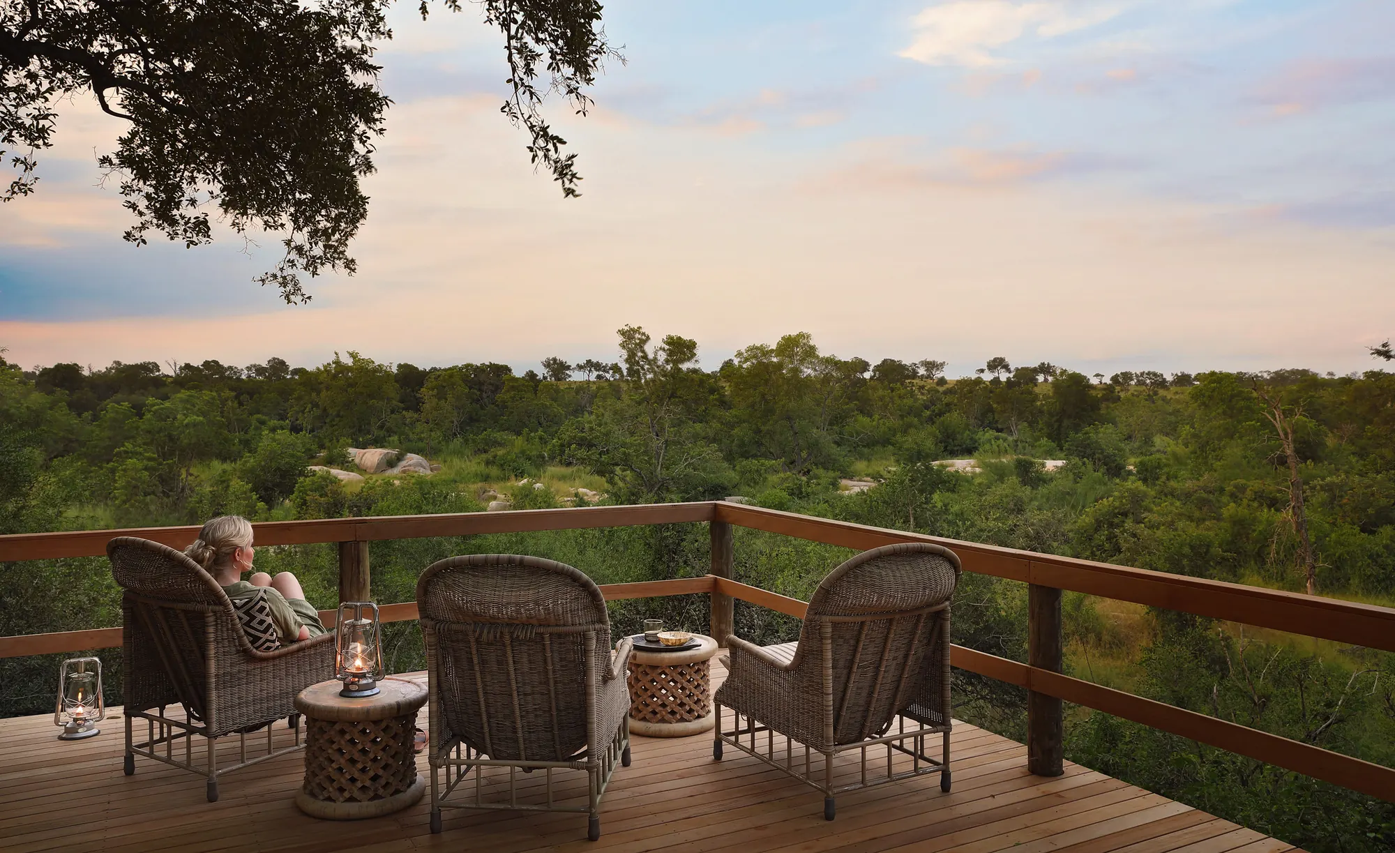 The main deck at Pioneer Camp at dusk — four woven armchairs and two lantern-lit side tables on raised timber, looking out over unbroken bush under a pale sky.