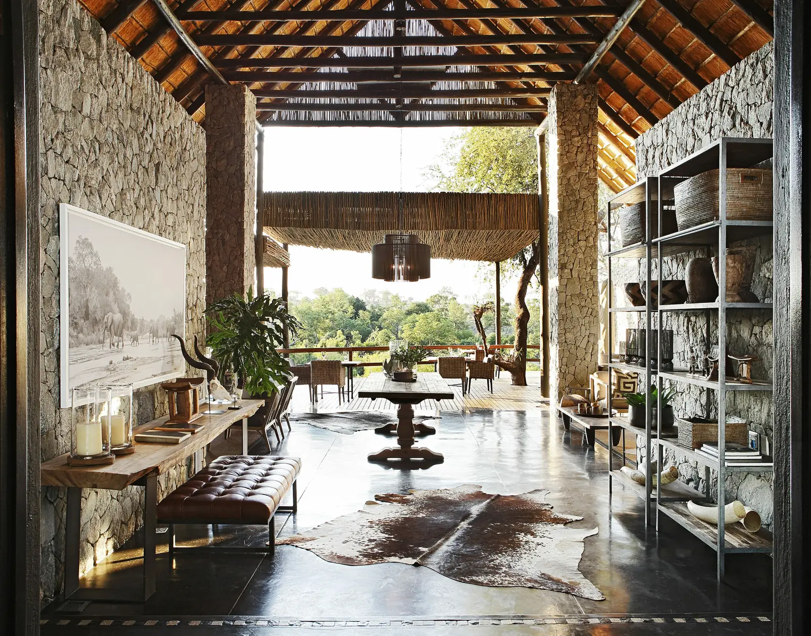 The stone-walled entrance hall of the Private Granite Suites under an exposed timber-and-thatch roof, with a candle-lined console and a tufted leather bench, looking through to a deck and trees beyond.