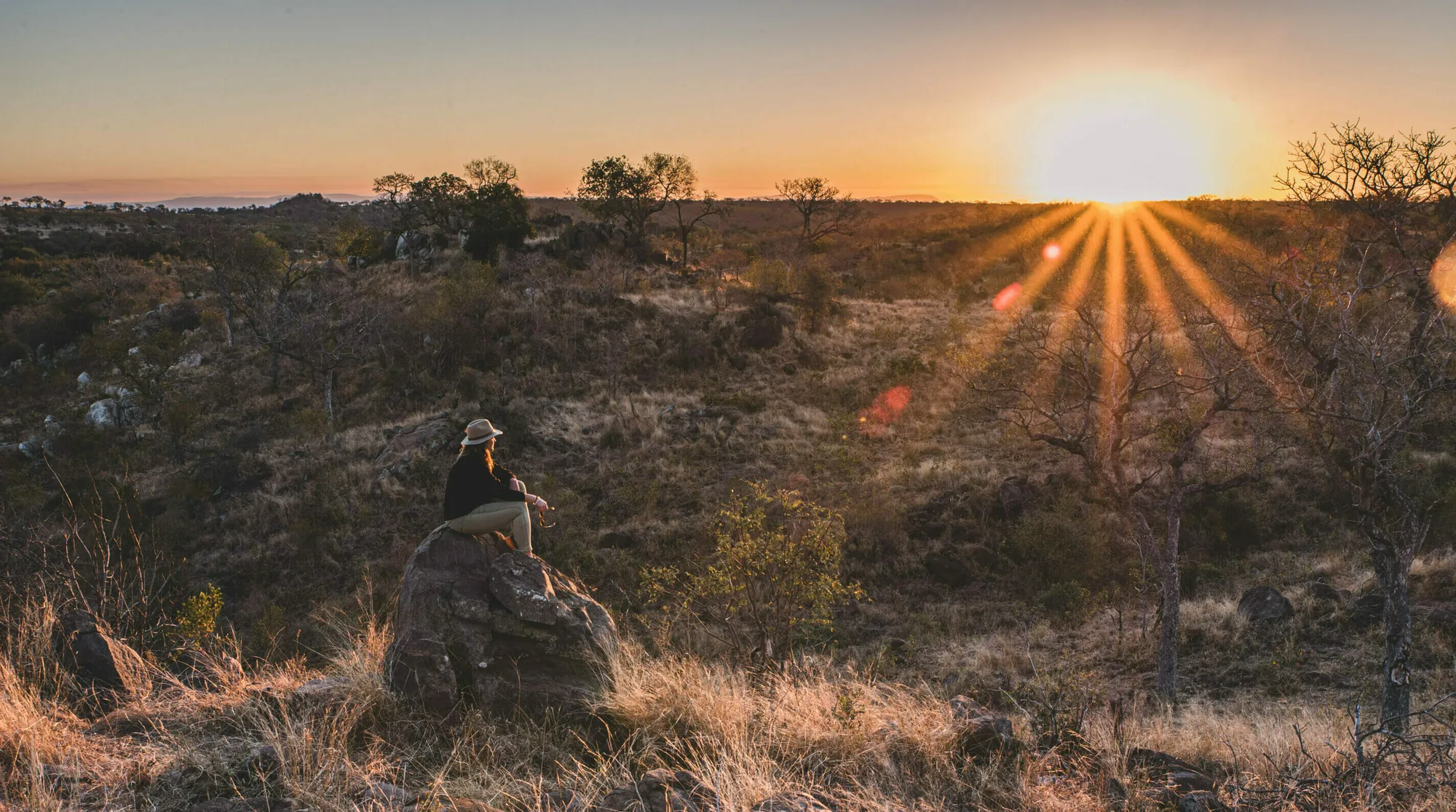 A lone figure seated on a rock in the middle distance, dry grass in the foreground, the sun low and flaring behind a wooded ridge.