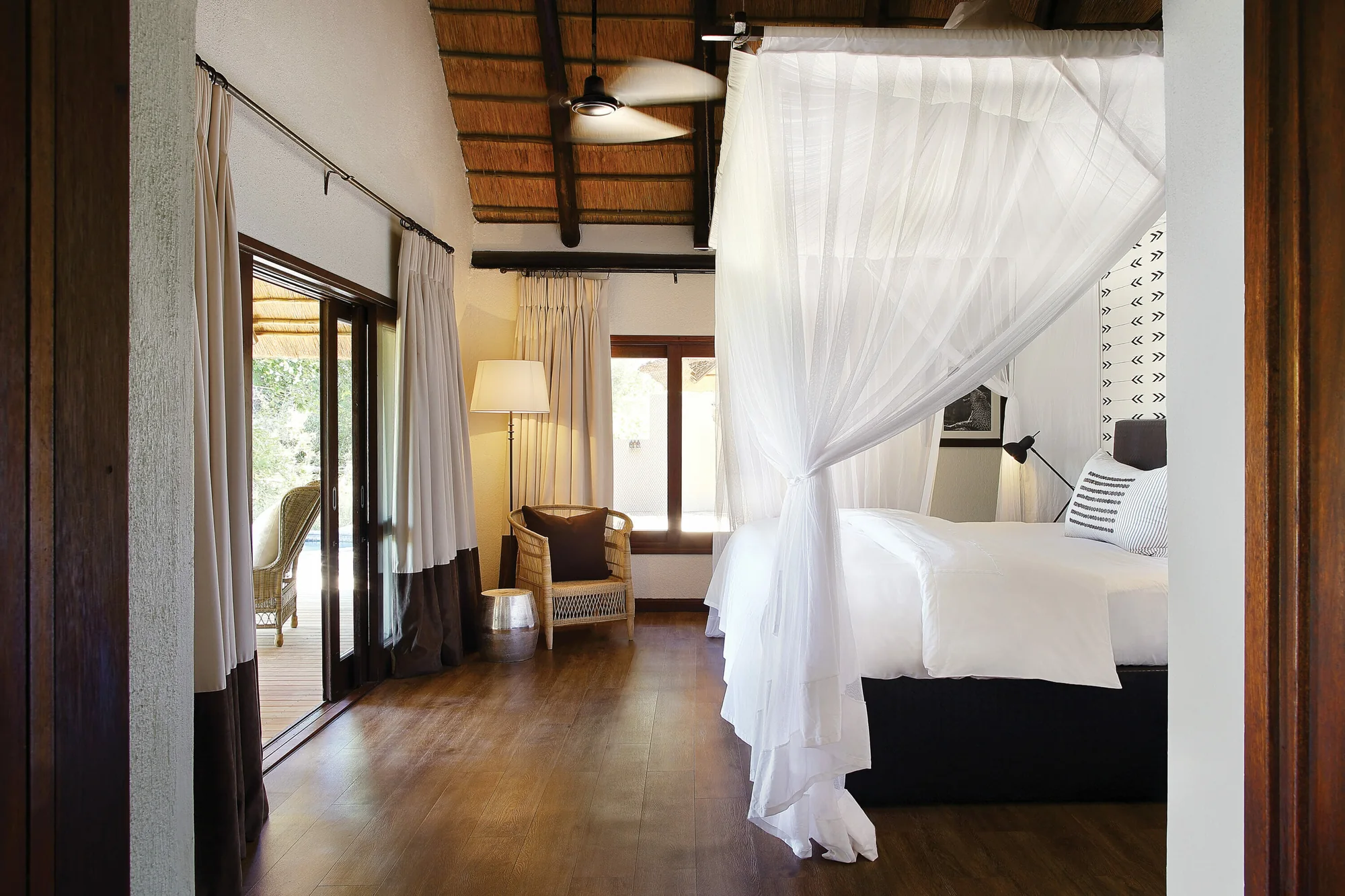 A bedroom in a Tree Camp suite under an exposed thatch ceiling, with a white draped canopy bed and sliding glass doors onto a deck.