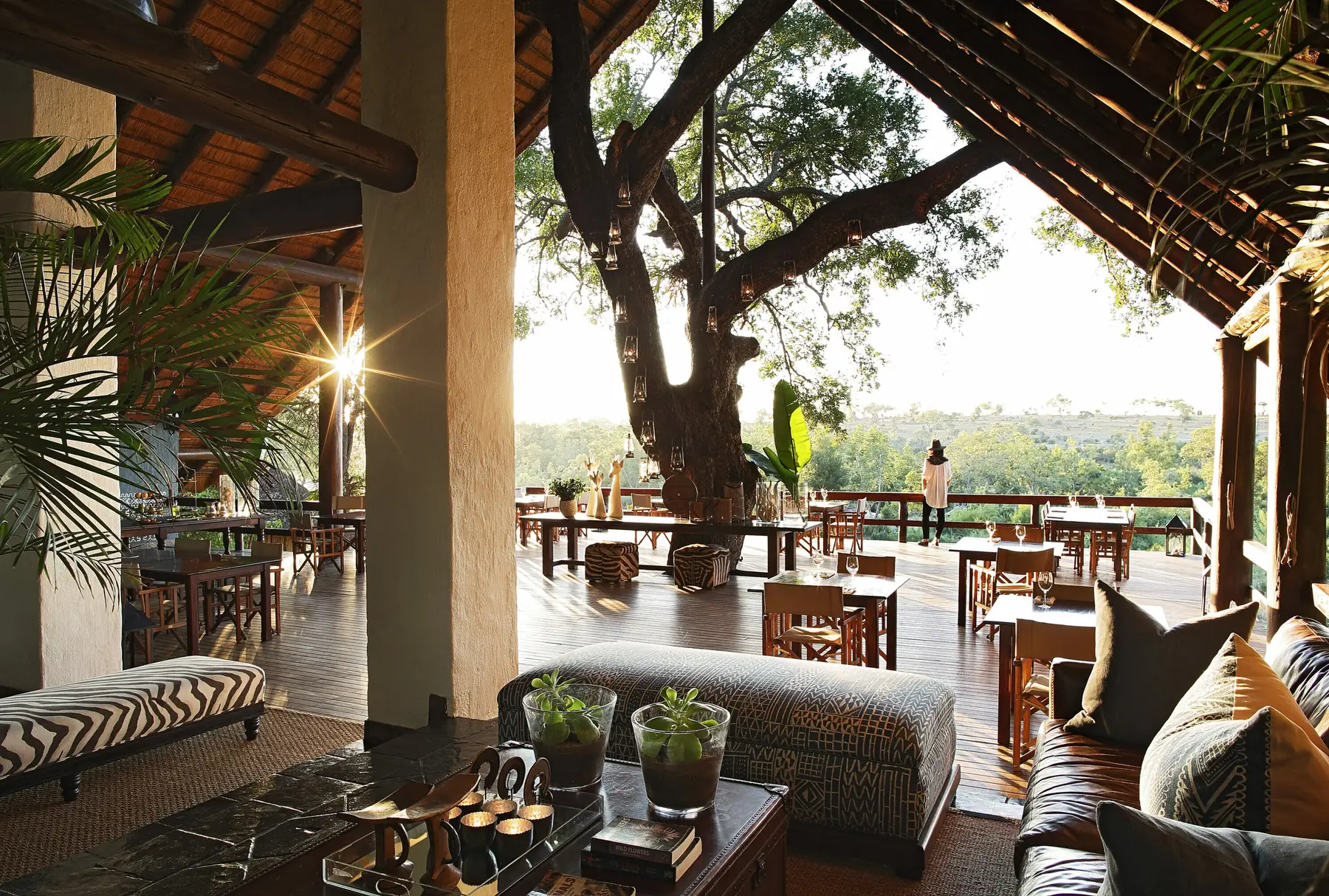 The open-sided main lounge at Varty Camp — leather sofa, zebra-hide bench and low table — opening onto a dining deck of small tables beneath a large tree, with bush and low sun beyond.